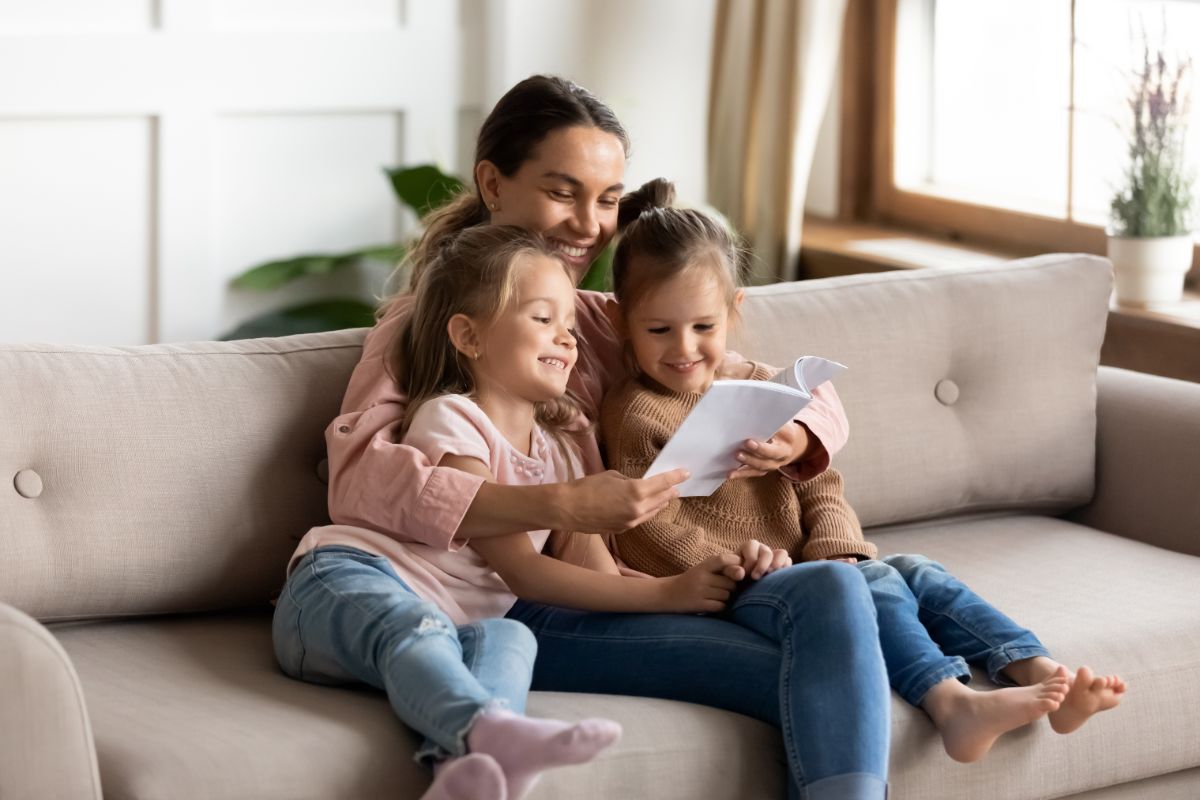 Young mother and her two daughters sitting on a couch and reading a book.