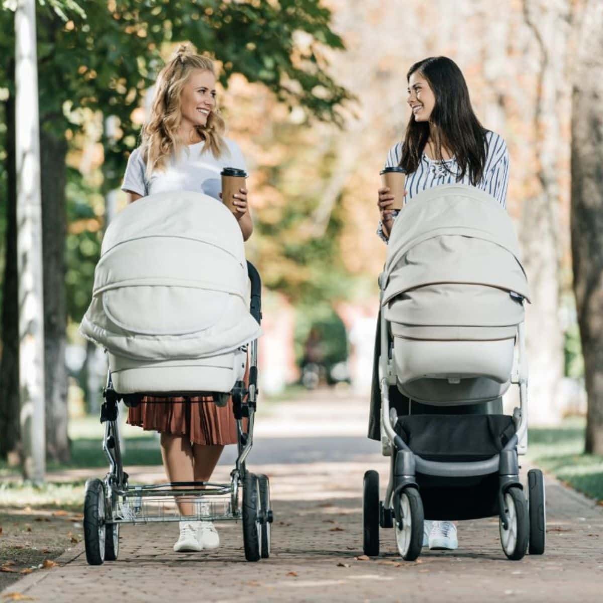 Two young mom pushing baby strollers in a park and talk to each other.
