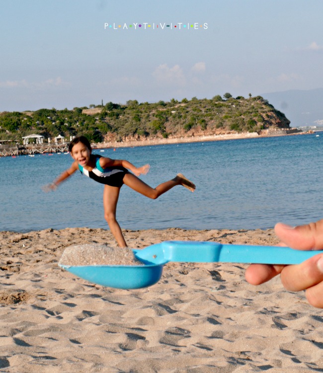 Hand holding a blue spoon with a sand and little girl standing on a beach.