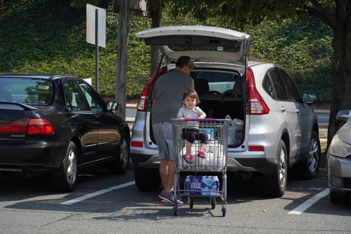 Father picking up purchased items from a shopping cart into a car while his daughter sitting in a shopping cart.