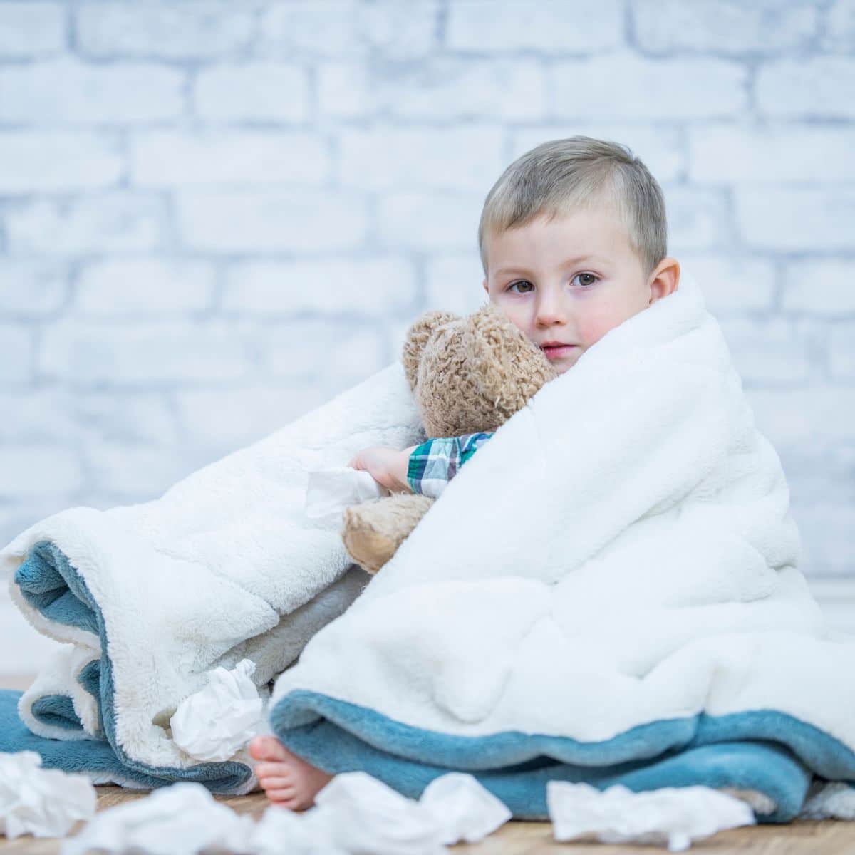 Sick looking toddler covered in a blanket holding a stuffed toy and napkins around him.