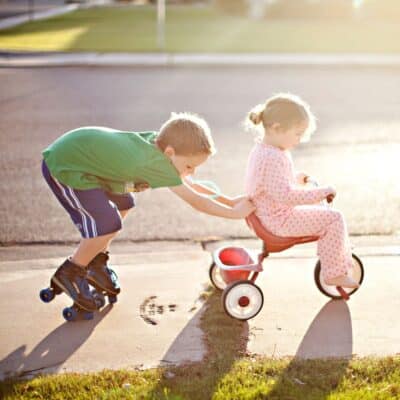 Brother on roller skates pushing younger sister on a tricycle.