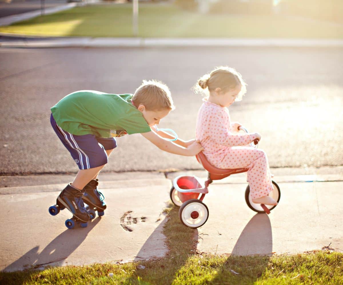Brother on roller skates pushing  younger sister on a tricycle.