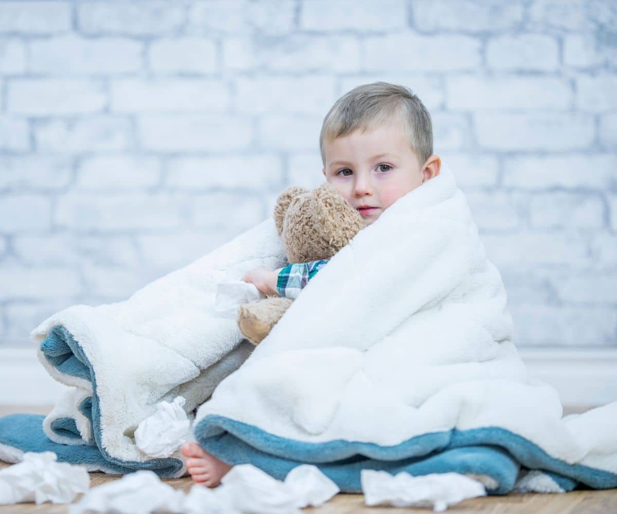 Sick looking toddler covered in a blanket, holding a stufffed bear and napkins aroung him.