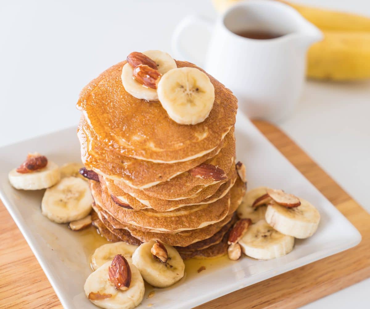 Pile of yummy looking pancakes with sliced banana and nuts on the top and on a plate.