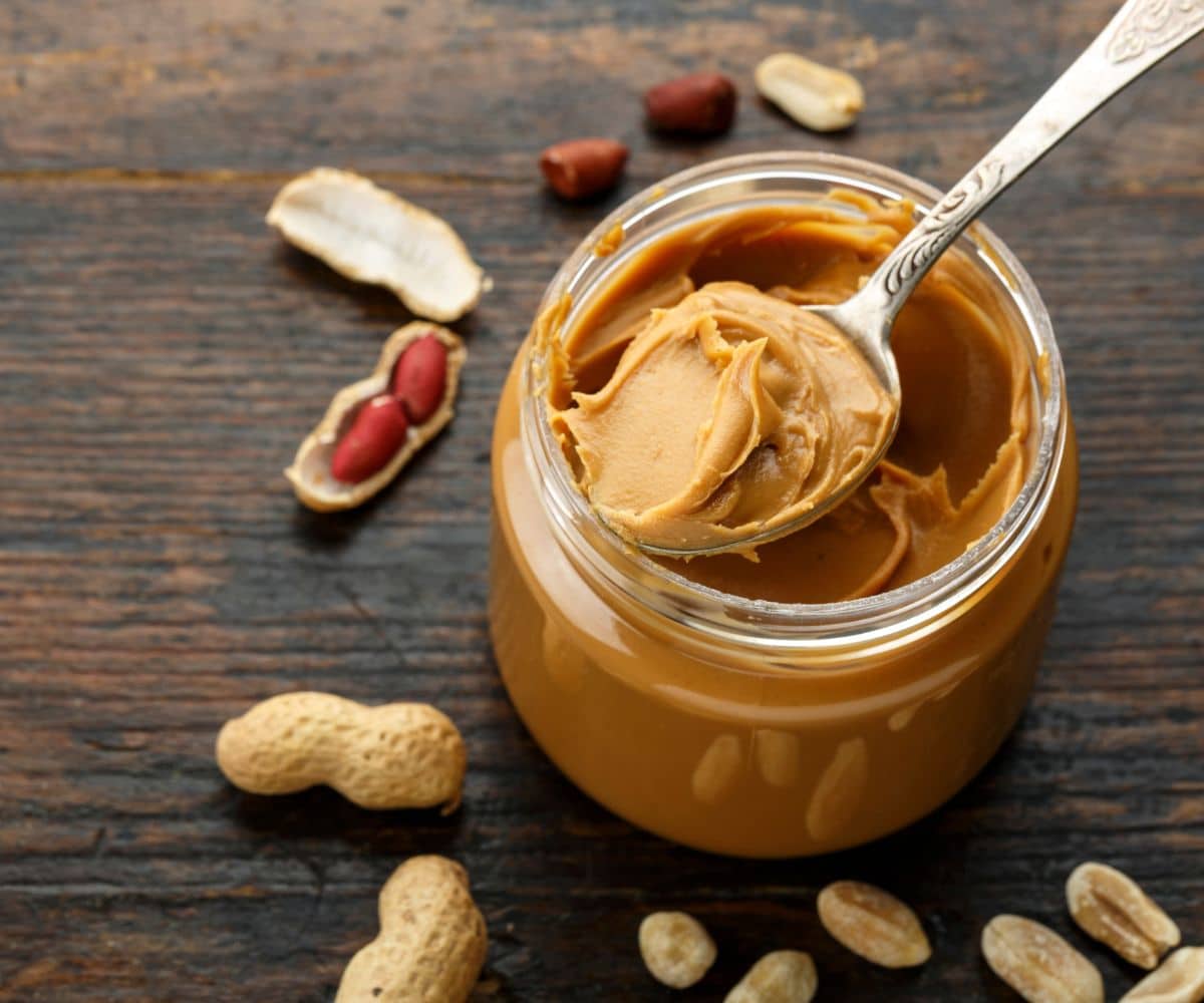 Jar full of peanut butter with a spoon on a table with scattered peanuts around.