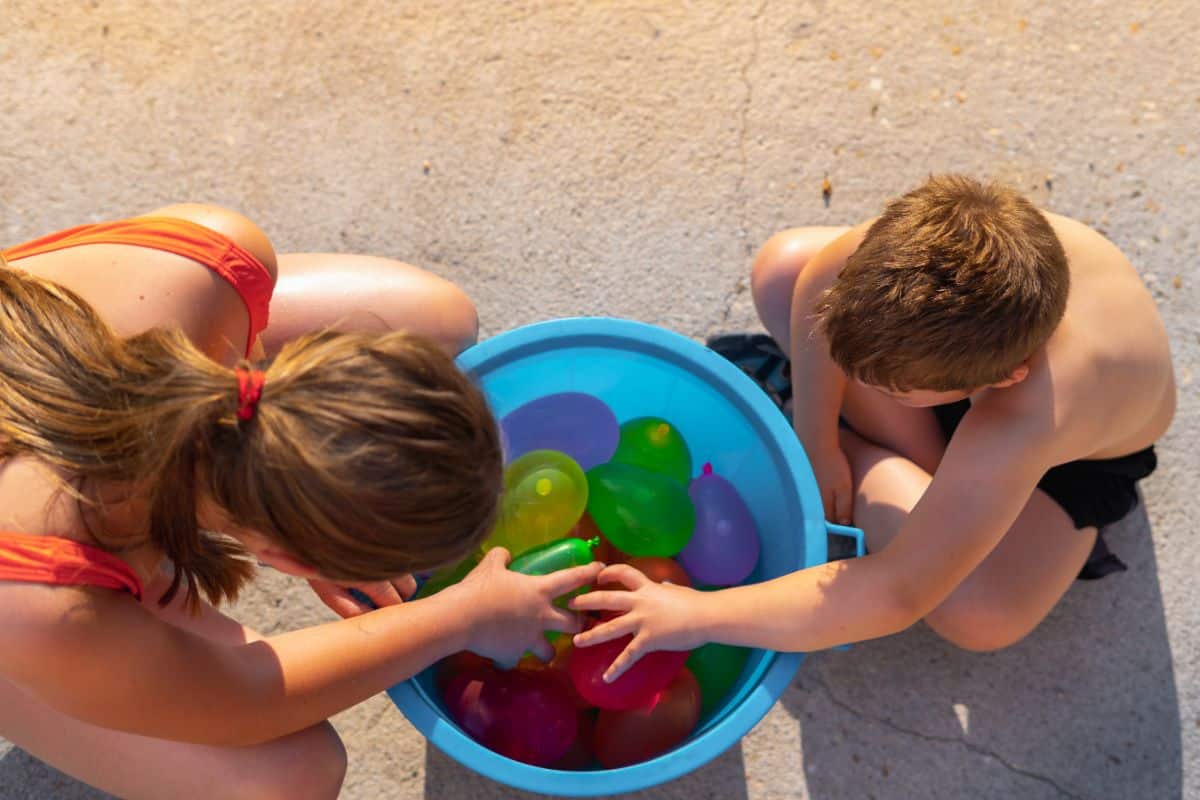 Two kids picking up water balloons from a blue bowl.