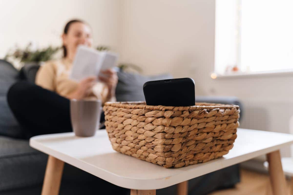 Mobile phone in a basket on a table while woman reading a book in the background.