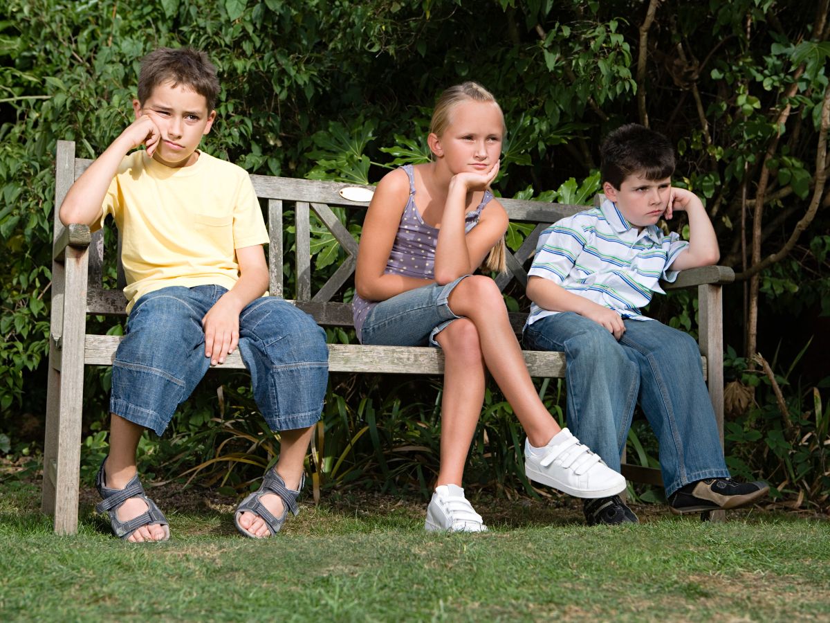 Three kids sitting on a wooden bench outdoor looking bored.
