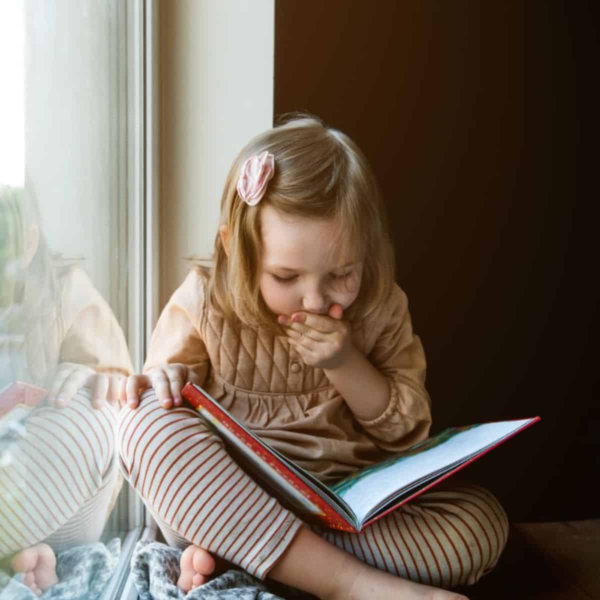 Young little girl sitting near a window and reading a book.