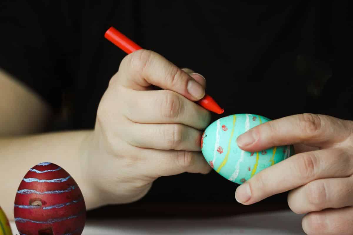 Hands painting with a red wax crayon an easter egg.