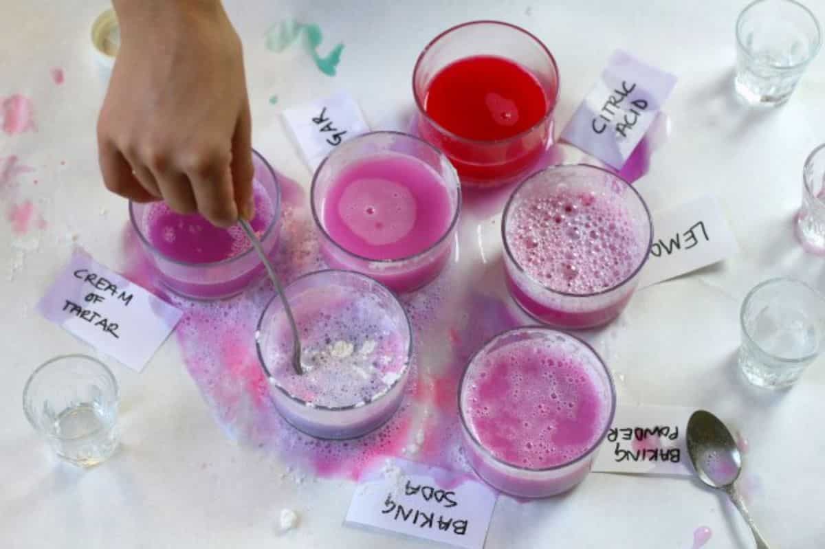 Hand is stirring a juice in a glass cup on a table with other glass cups of juice on a table with a spoon and stickers.
