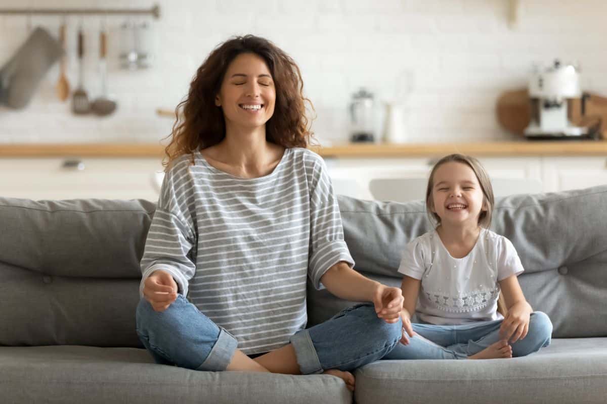 Smiling mother and daughter meditating on a gray sofa.