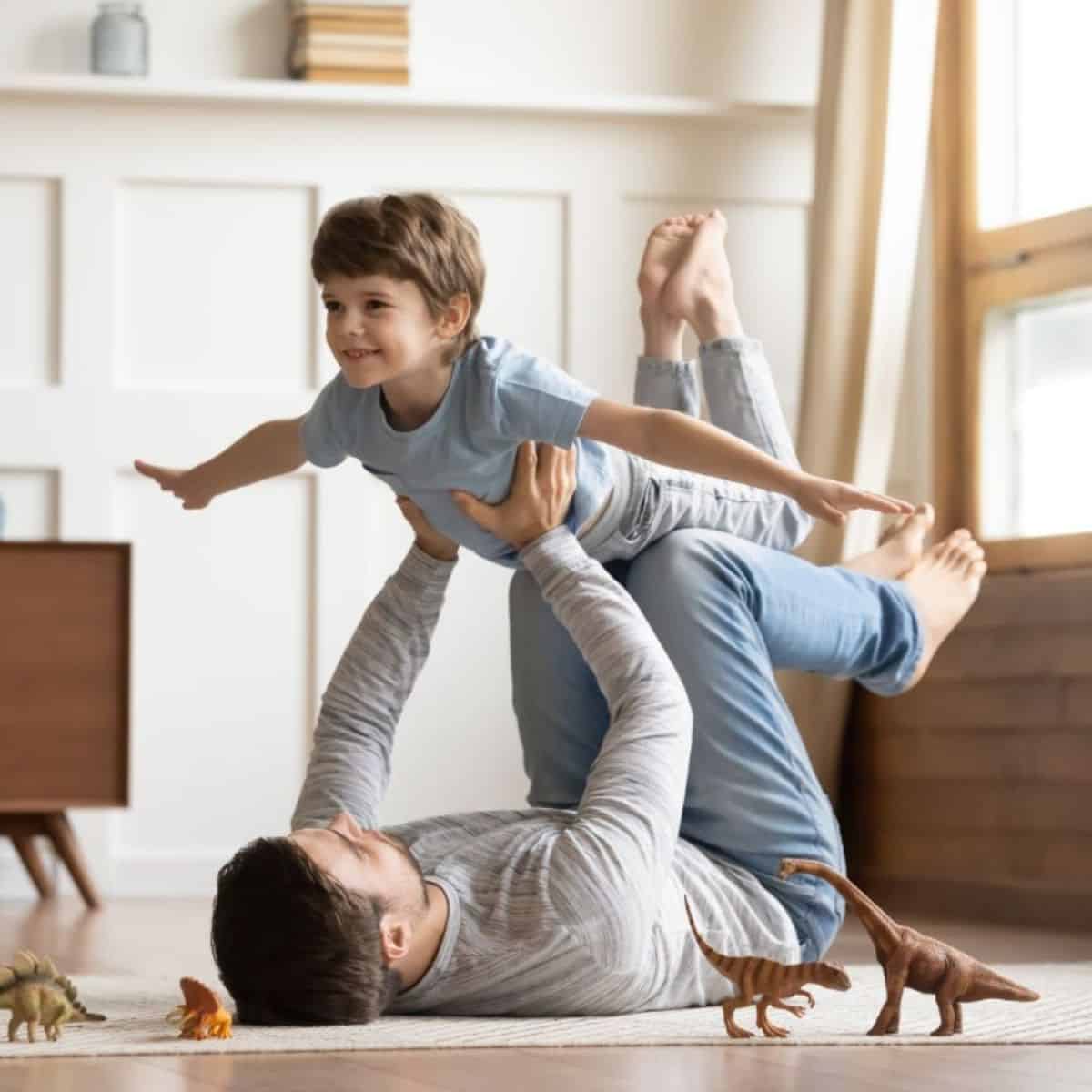 Young father holding his son in air by hands and feet in a living room.