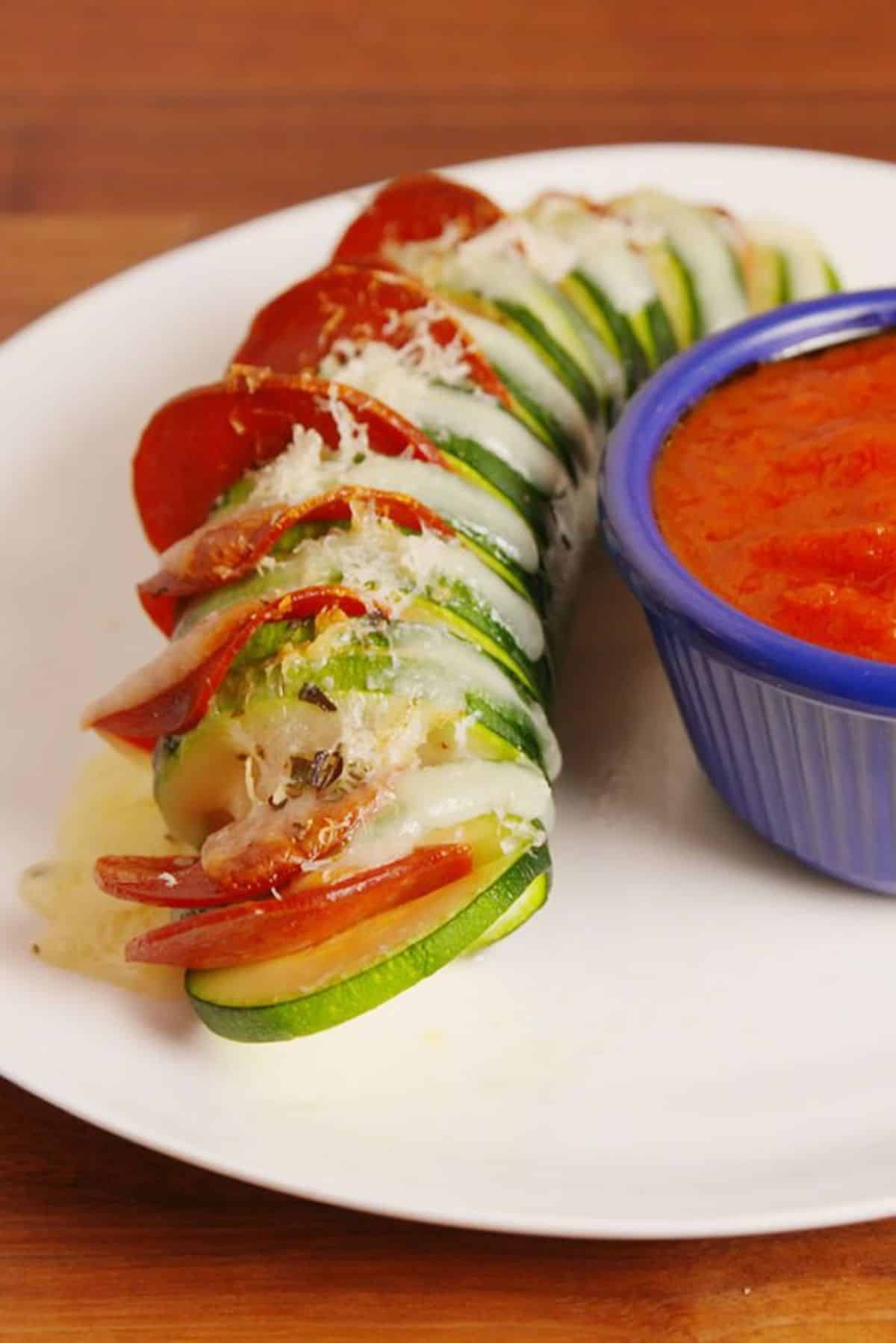 Pizza Stuffed Zucchini on a white plate with bowlf of sauce.
