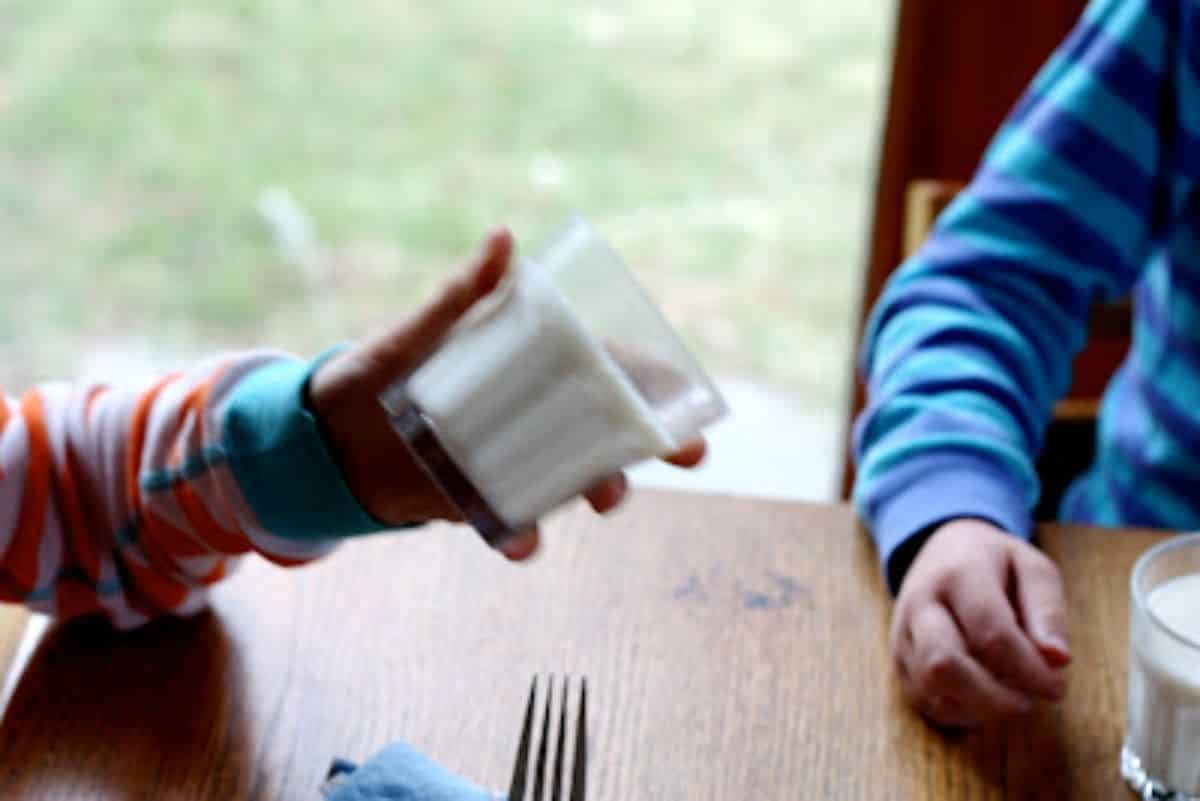 Kid hand holding a fake glass of milk towards another kid sitting at a table.