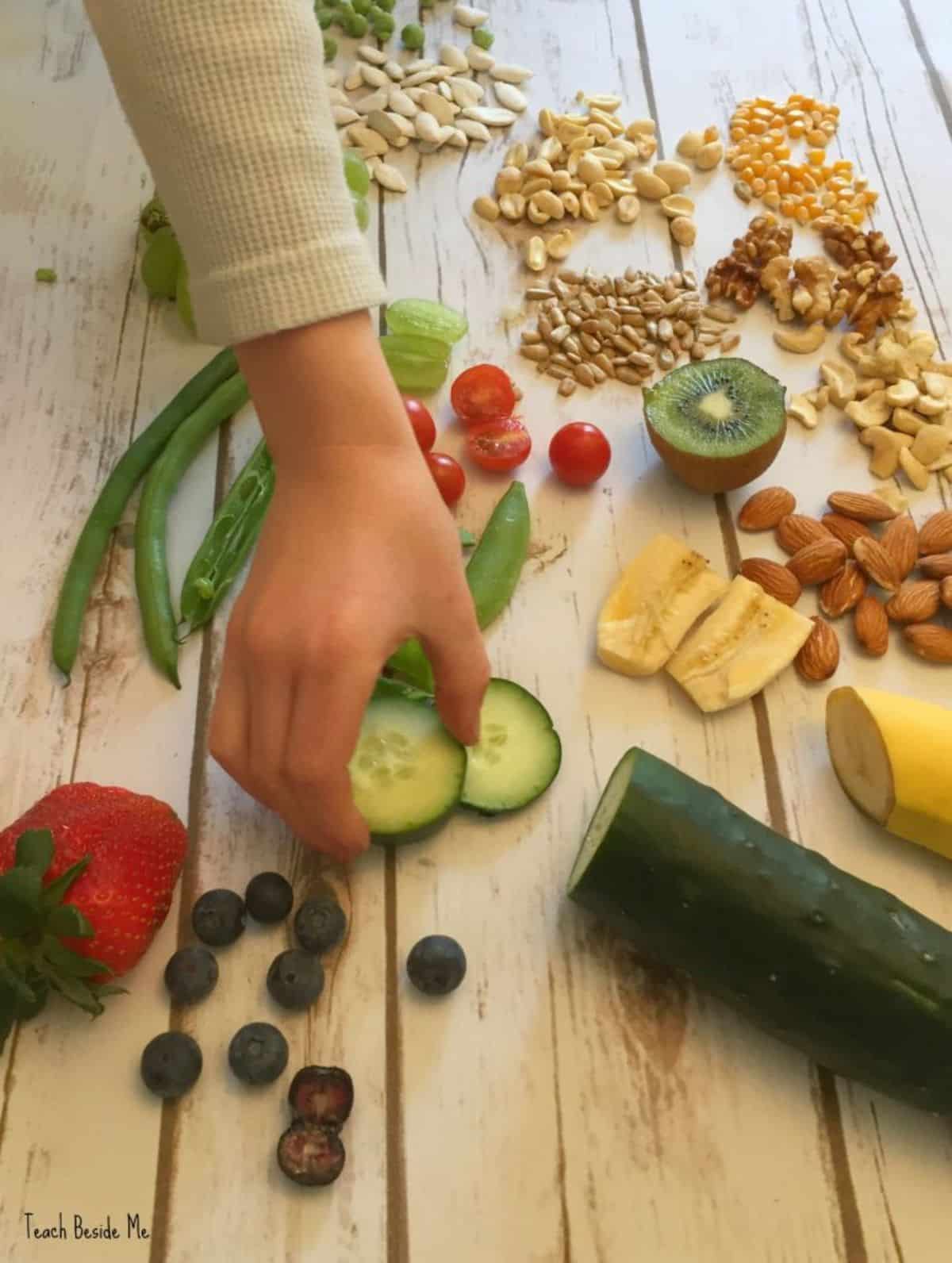 Hands are touching slices of cucumber on a wooden table with sliced vegetables, fruits and nuts.
