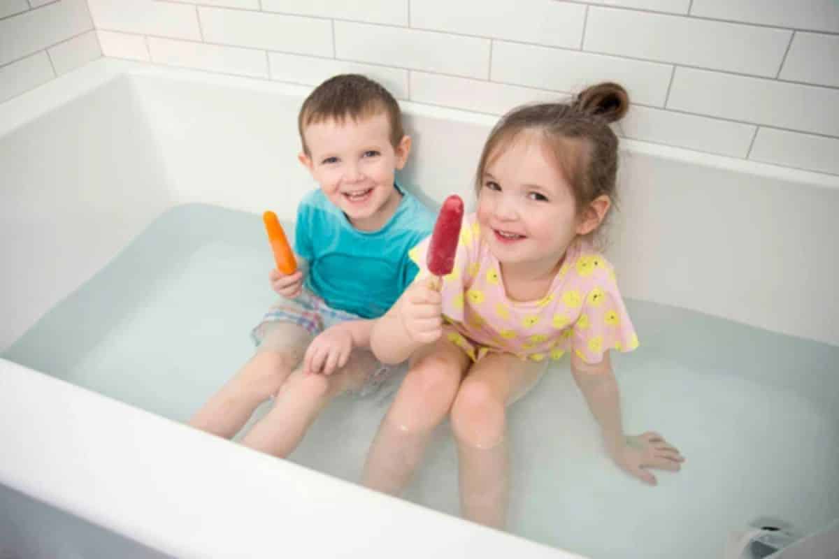Two kids with holding popsicles in a bathtub with water.