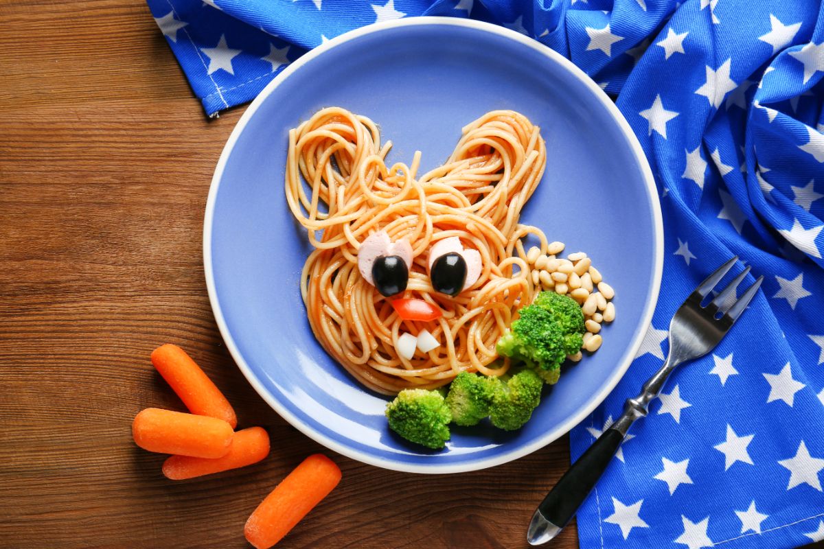 Pasta designed as a face with vegetables on a blue plate on a table with a fork and small carrots.