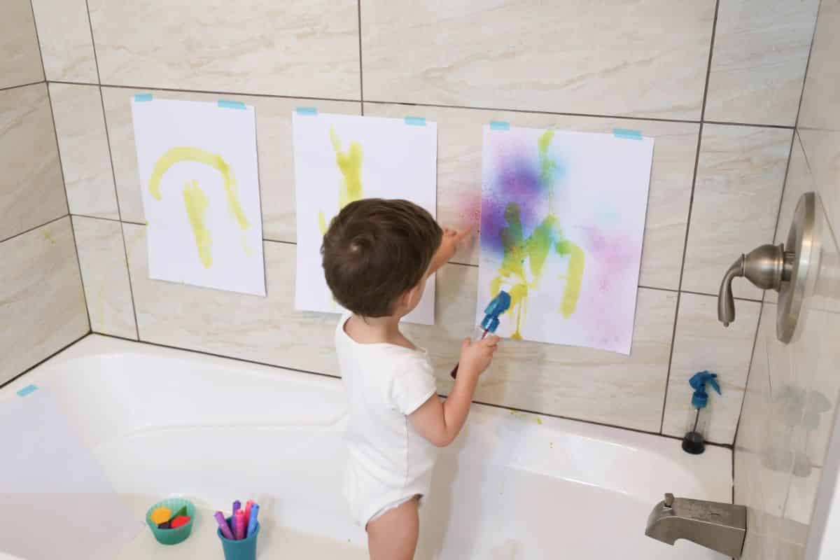 Small boy in the  bathtub painting a paper sheets hanging on the wall.