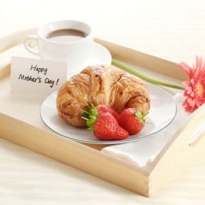 Croissant with ripe strawberries on a white palte on a wooden tray with a cup of coofe, a flower and sign.