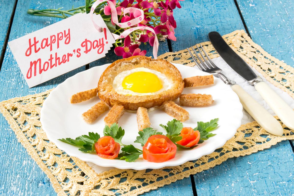 Bread with and egg, herb and sliced tomato on a white palte on a blue table with cutlery and bouquet of flowers with sign Happy Mother's day!
