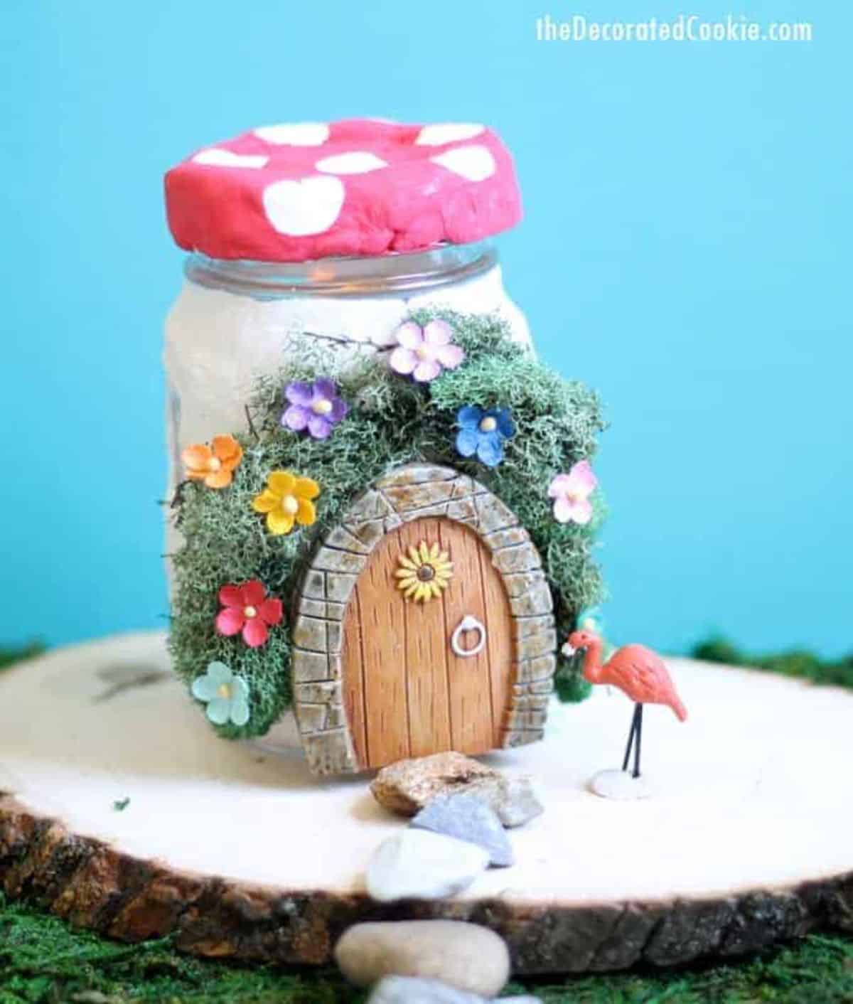 Mason jar fairy house on a wooden board with a tiny pink flamingo.