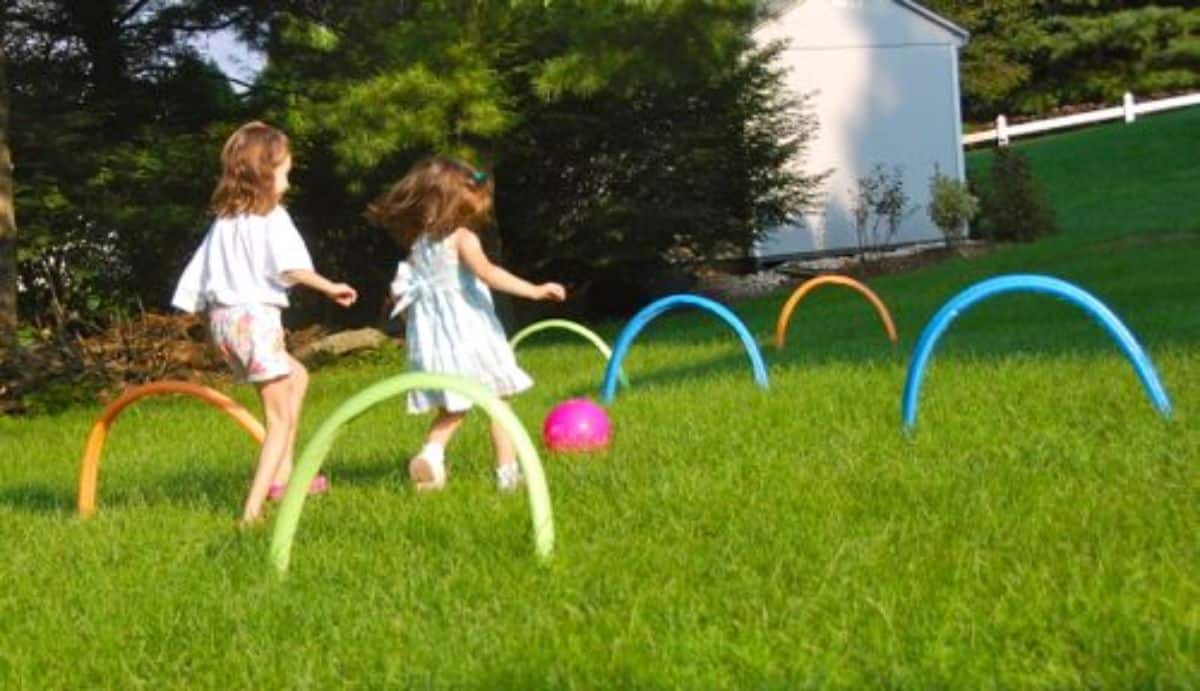 Two small girls playing on a green grass with old pool noodles sticked in the ground.