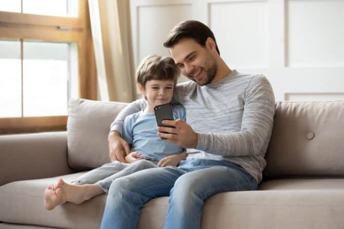 Father and son sitting on a gray sofa and watching something on a smartphone.