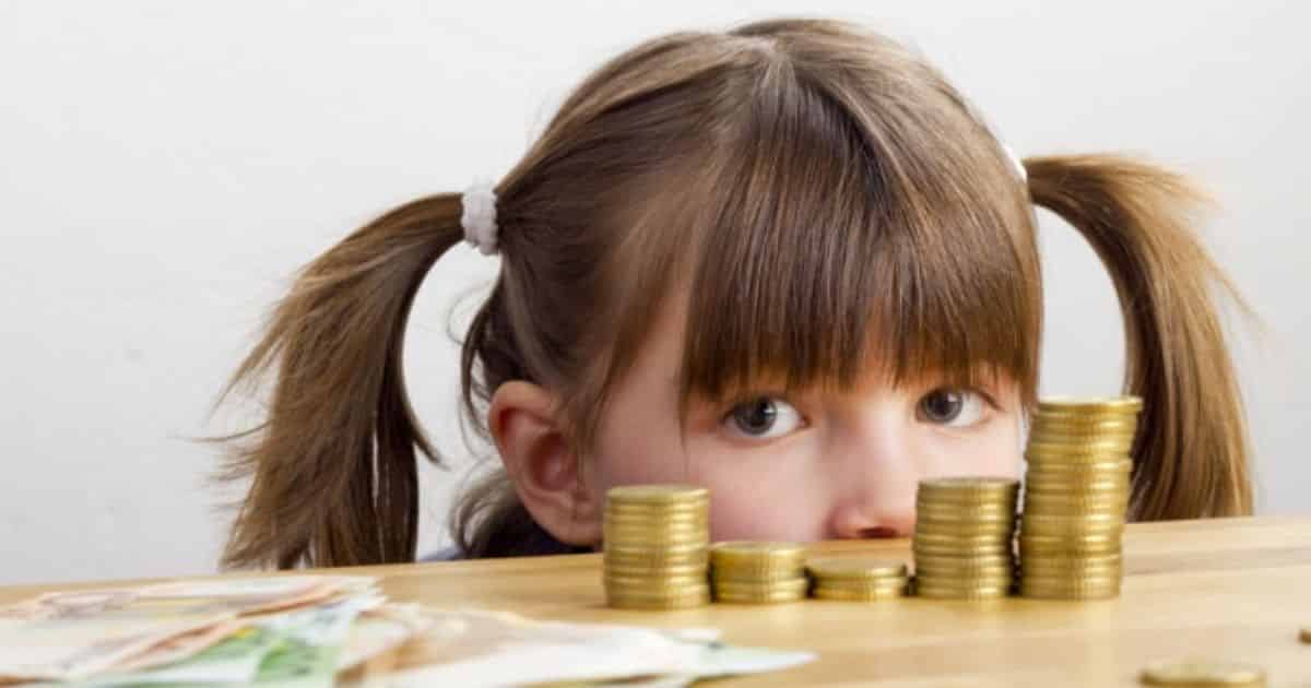 Little girl with ponytails behind the table looking at stash of coins on the table.