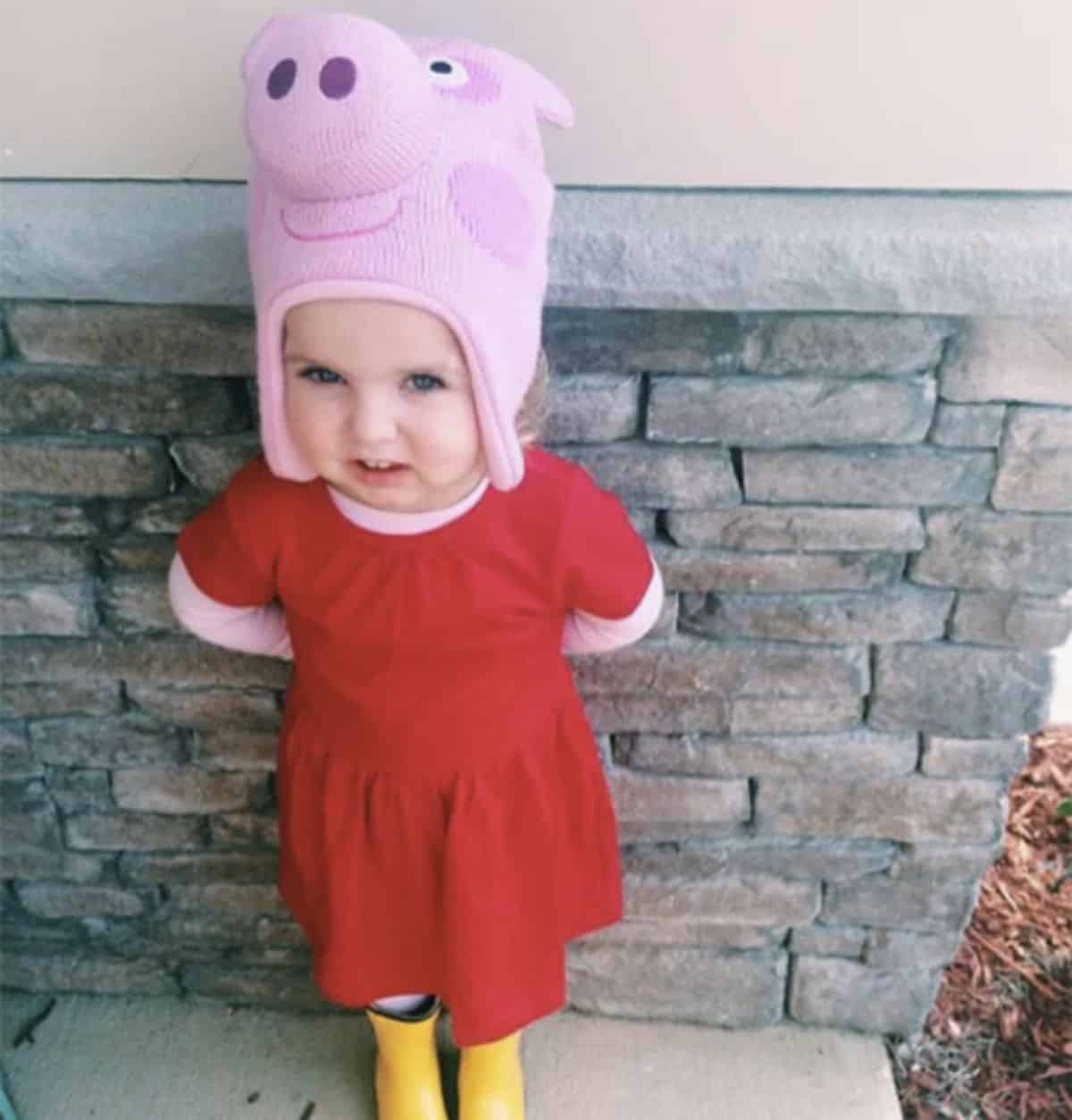 Little girlin a red dress with peppa pig hat leaning on a wall.