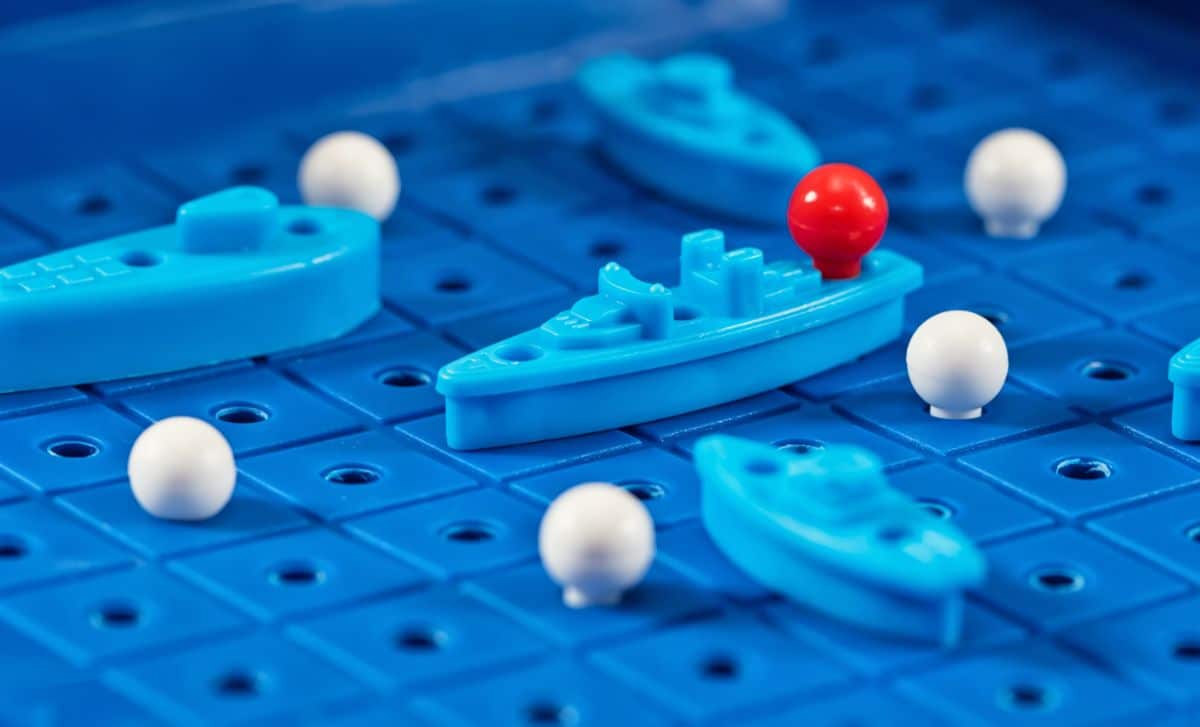 A close up of a battleship board with 4 blue boats, 5 white pegs and 1 red peg sitting in the bow of a ship