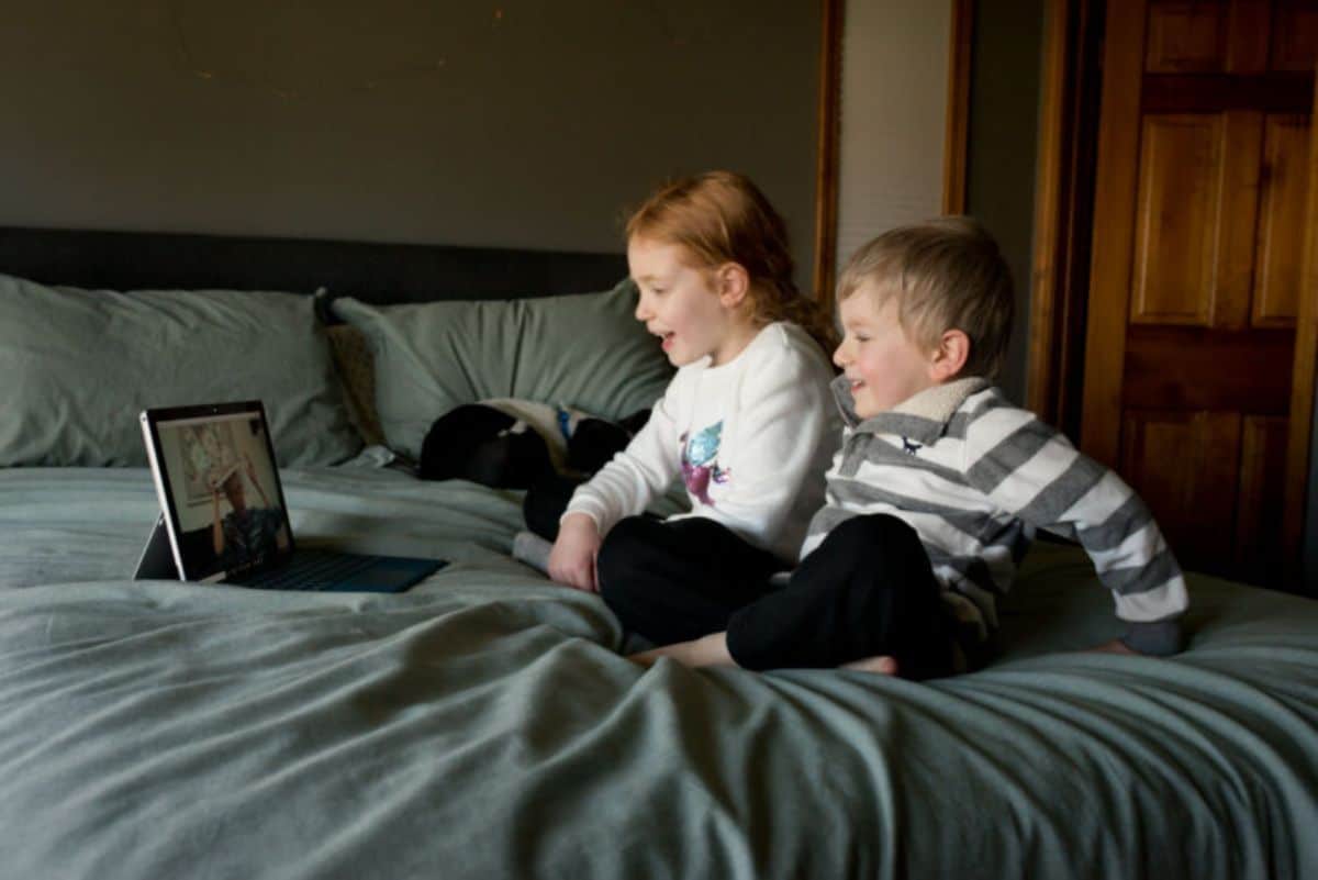 2 children sit cross-legged on a bed looking at a tablet propped up.