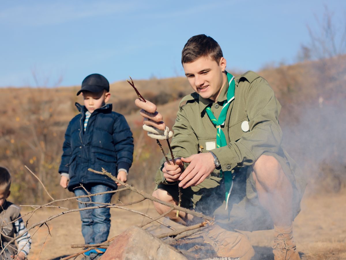 A scout is crouched before a fire holding dough on twigs. A child in a coat is standing behind him