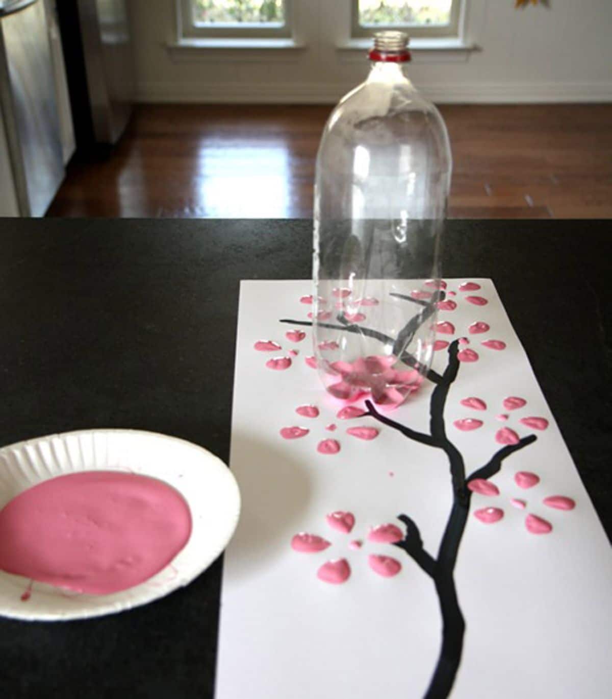 on a table is a paper plate full of pink paint, a white piece of paper with a black branch painted on it, and paint petals. A 2 litre water bottle sits on the paper with paint on the bottom