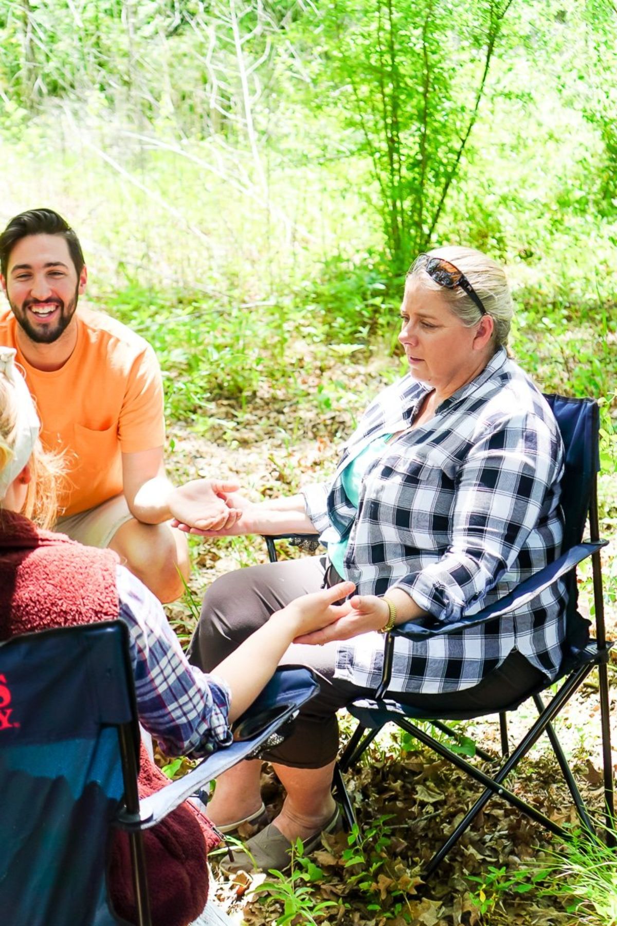 a group of 3 adults are sitting in a forest on camping chairs, holding hands