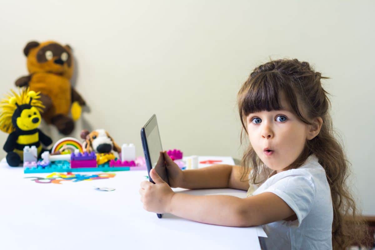 a youjng girl sits at a table holding onto a tablet. She is looking at the camera with a srprised expression on her face. In the background are some soft toys and lego