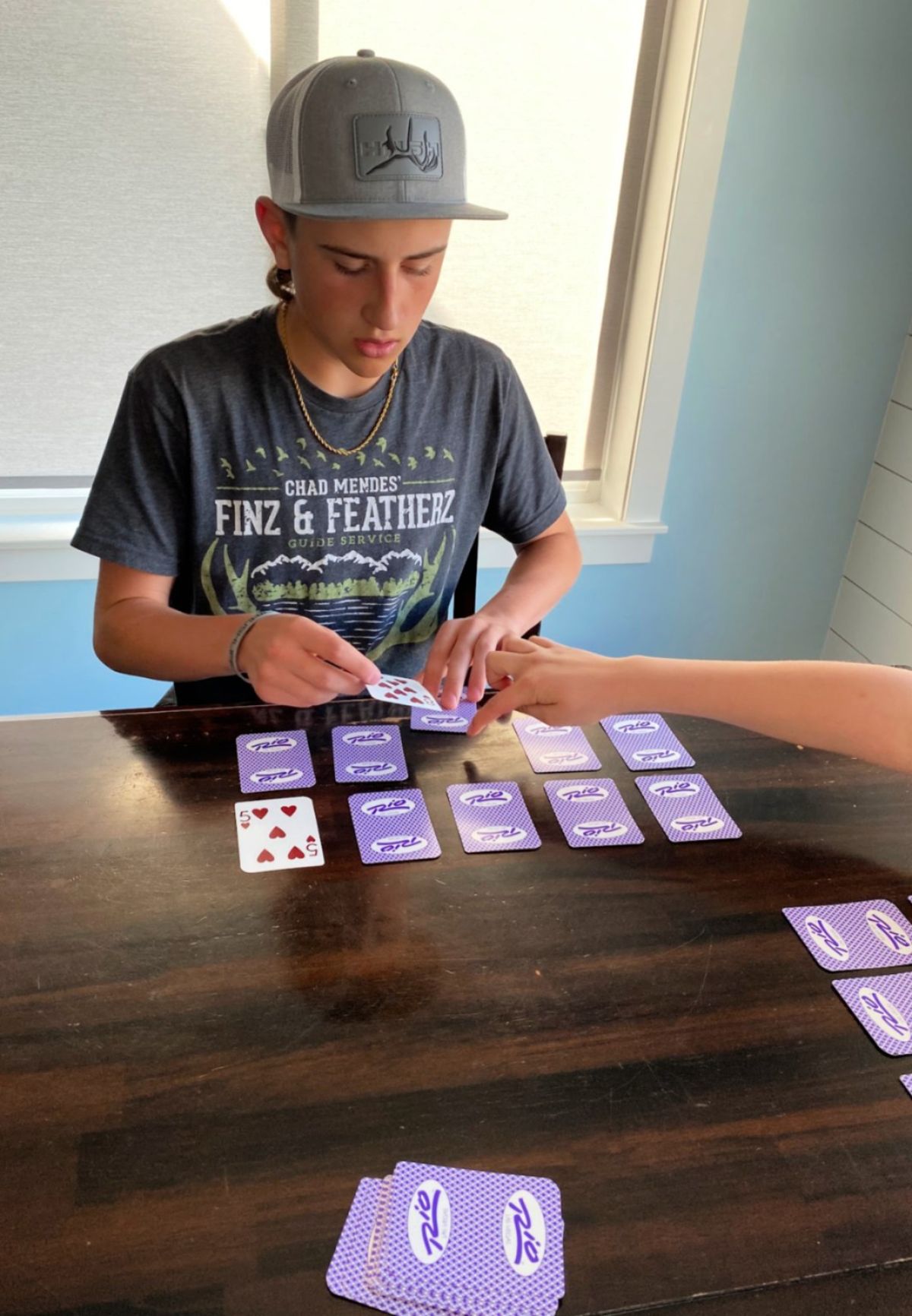 a boy sits at a dark wooden table and deals playing cards with purple backs