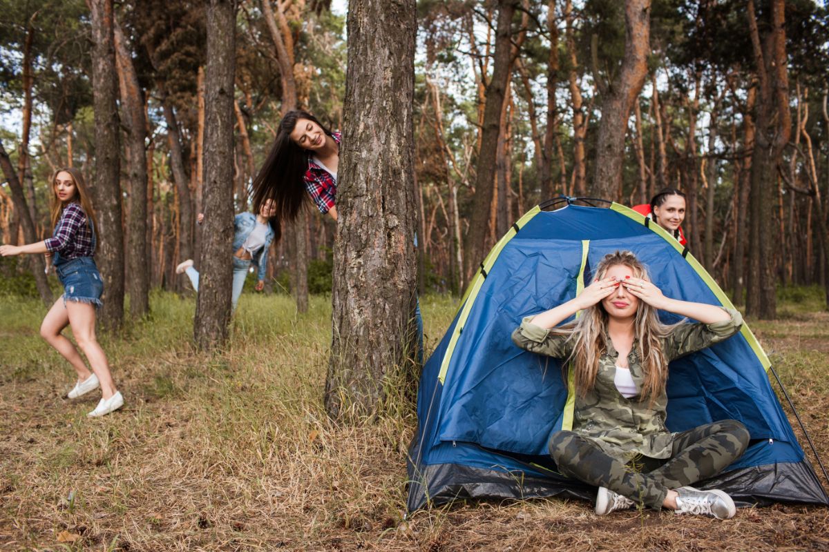 a girl sits in front of a blue pop-up tent with her hands over her eyes. 4 other girls are hiding behind trees smiling