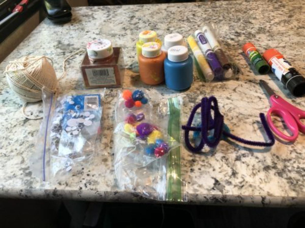 on a kitchen counter is a collection of craft materials: some string, googly eyes in a bag, ink, paint pots, pens, glue, scissors, pipe cleaners and tiny pom poms