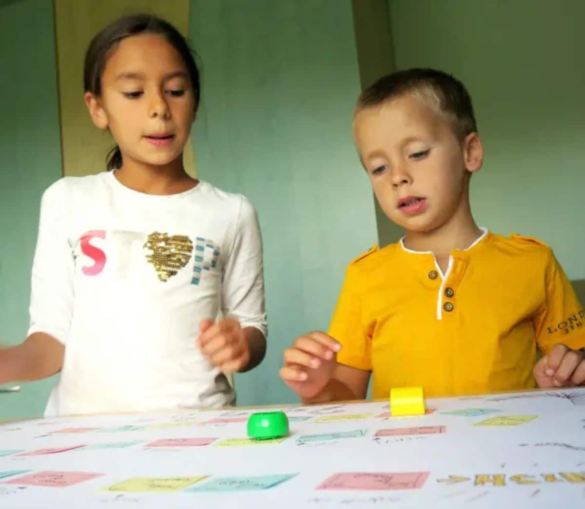 a boy and a girl look down at a board game with green and yellow counters on it