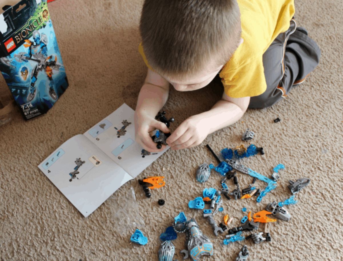 a child in a yellow shirt kneels on a carpetted floor putting together a lego model with a book of instrctions