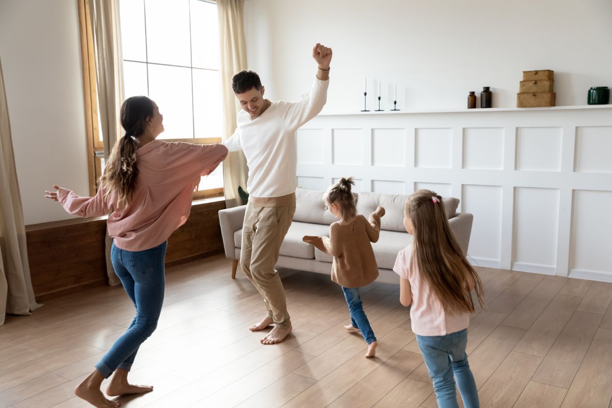 in a white living room with a wooden floor a family of four dance in front of a cream sofa