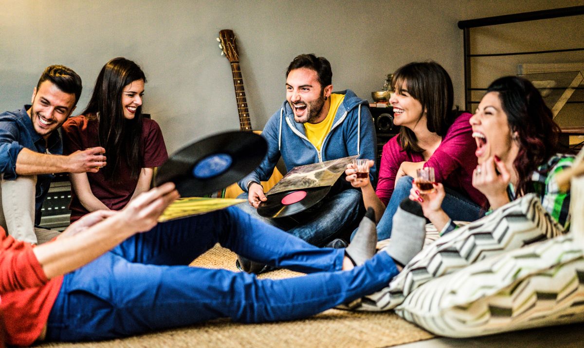 A group of youjng people sit on the floor of an apartment, holding records, glasses and laughing