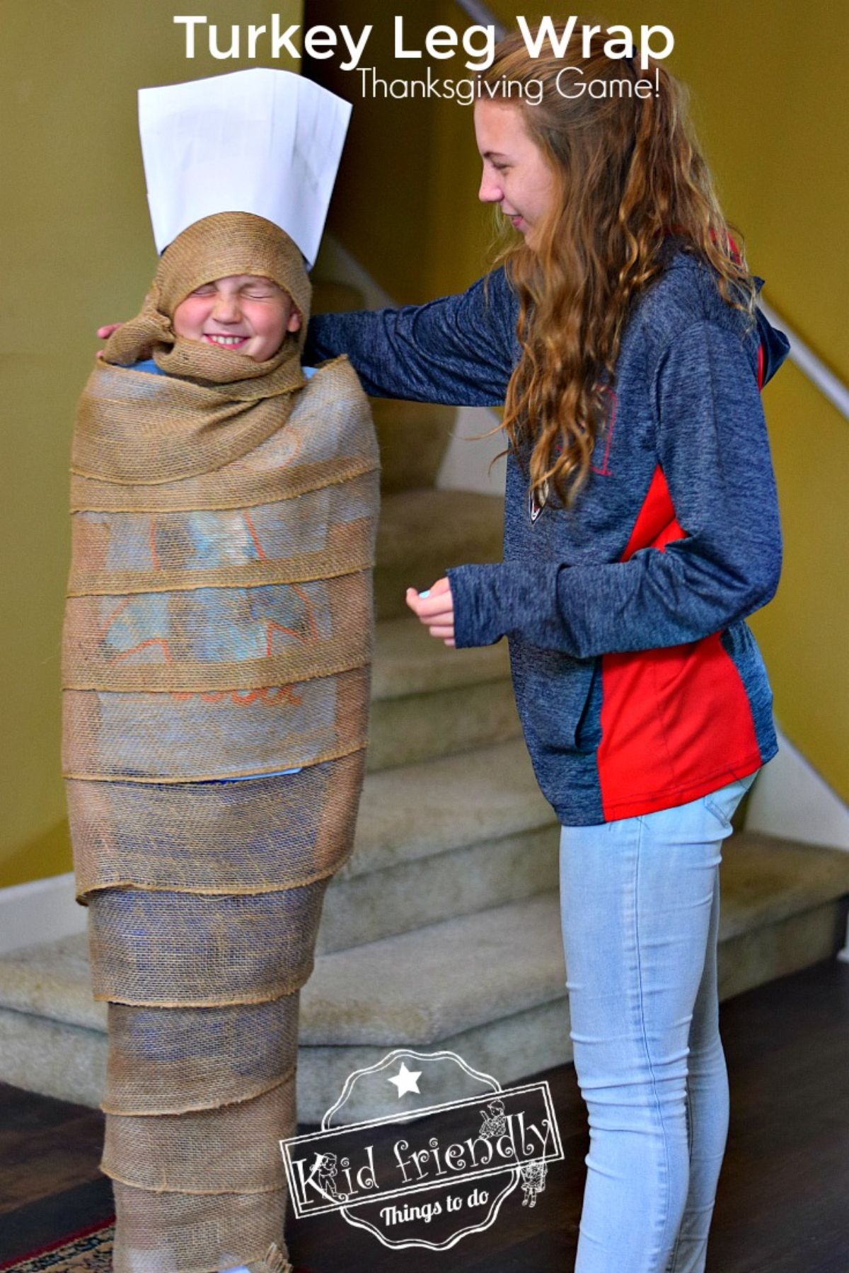 the text reads "turkey leg wrap thanksgiving game". The image is of two children, one wrapped in burlap with a chef hat on their head. The other is a girl with long hair tucking the burlap into his neck.