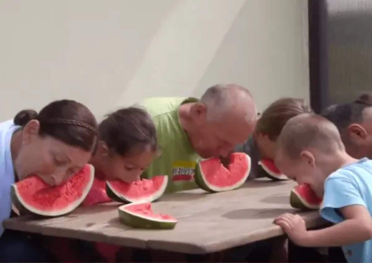 a man, woman and 3 children all bite into slices of watermelon sitting on a table