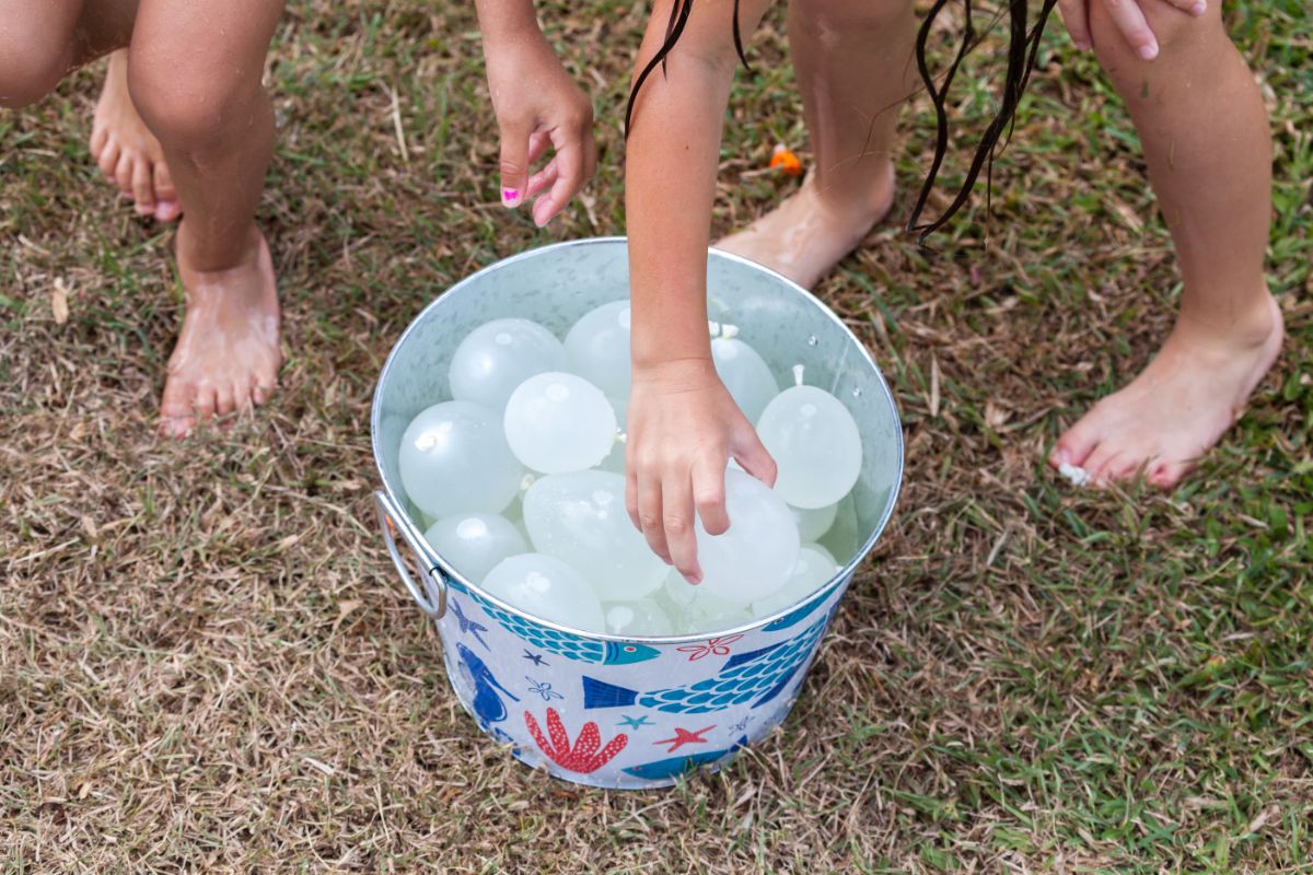 A metal bucket sits on the grass filled with water balloons. Hands are reaching into the bucket
