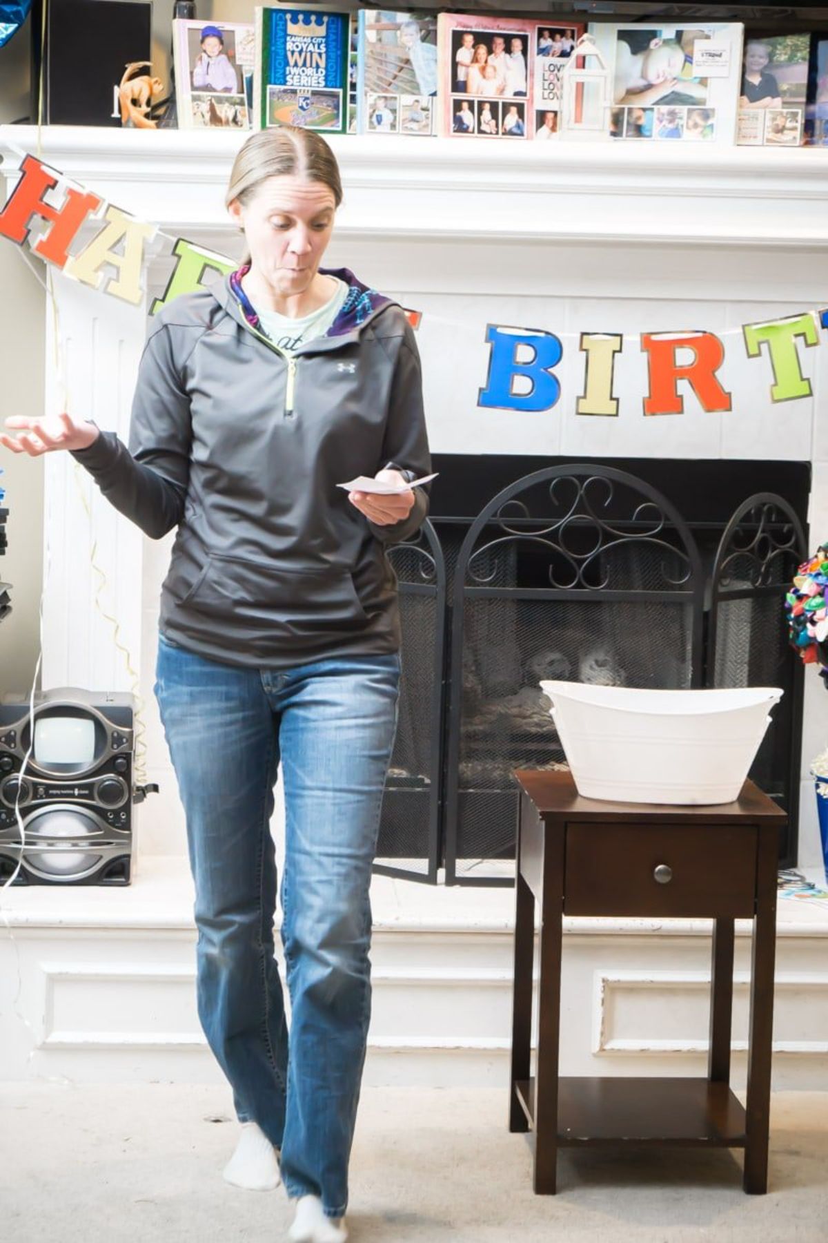 awoman is standing infront of a fireplace filled with birthday cards and draped with a happy birthday banner, She is holding a piece of paper