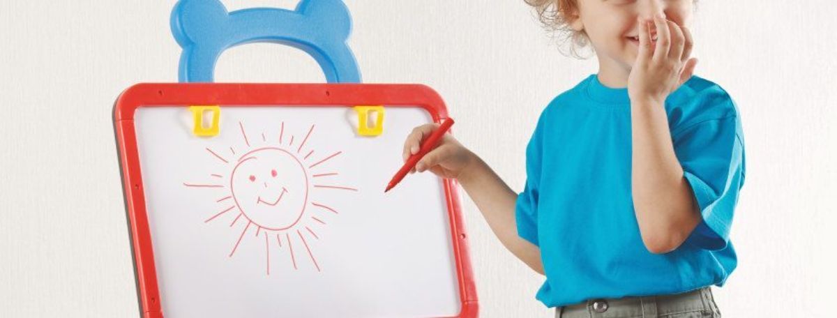 a small child in a blue shirt uses a red pen to write on a white board. She has drawn a sun with a smiley face