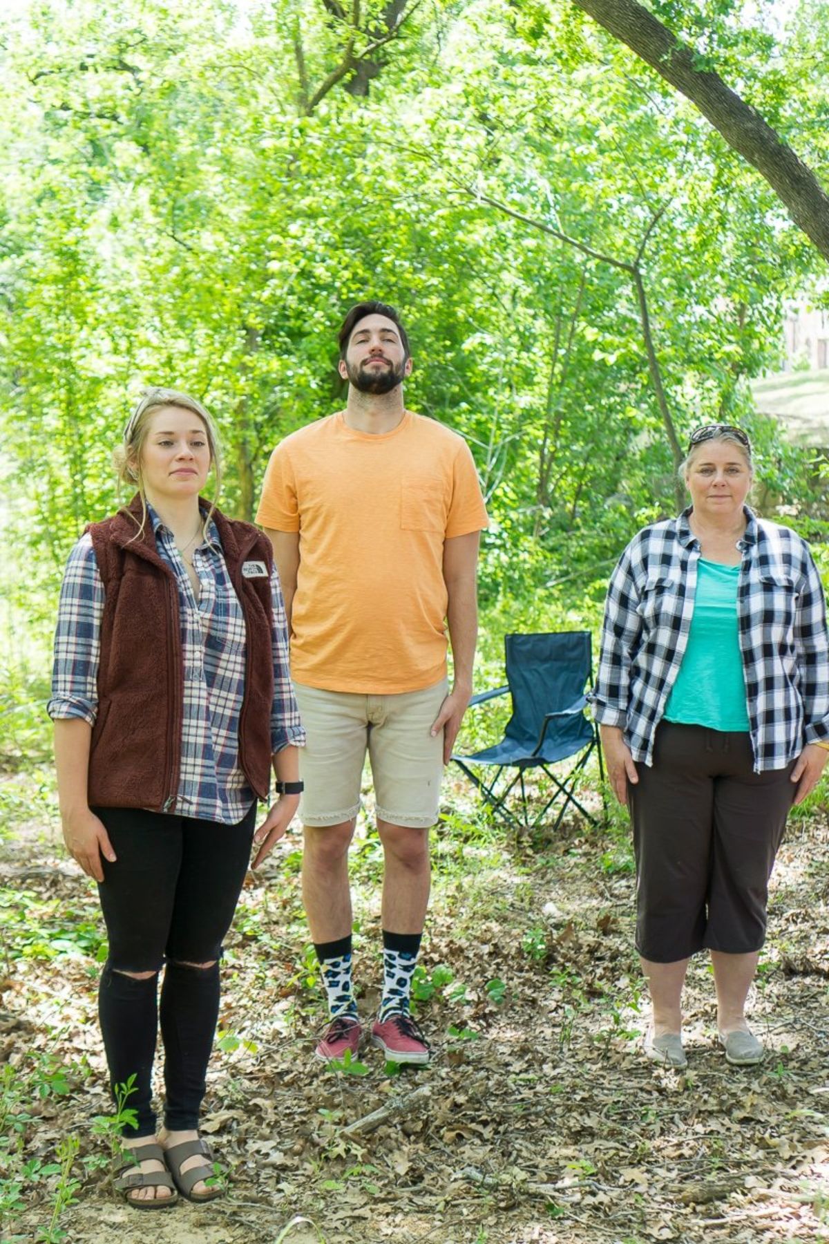 3 people stand in a wood with their hands by their sides. A camping chair is in the background